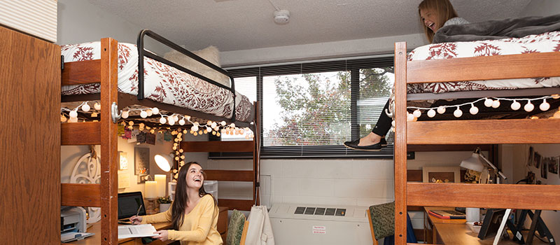 Furnished bedroom with 2 high beds with desks underneath. A young woman is at one of the desks and another is on one of the beds.