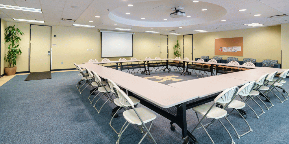 Activity room with long tables in the center in a square formation. Chairs around it, a wall screen, the GT logo in the center.