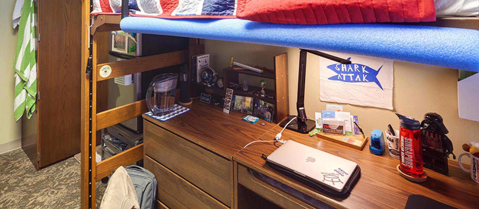 A desk under a bunk bed. THe desk has a laptop computer, desk lamp and other study items.