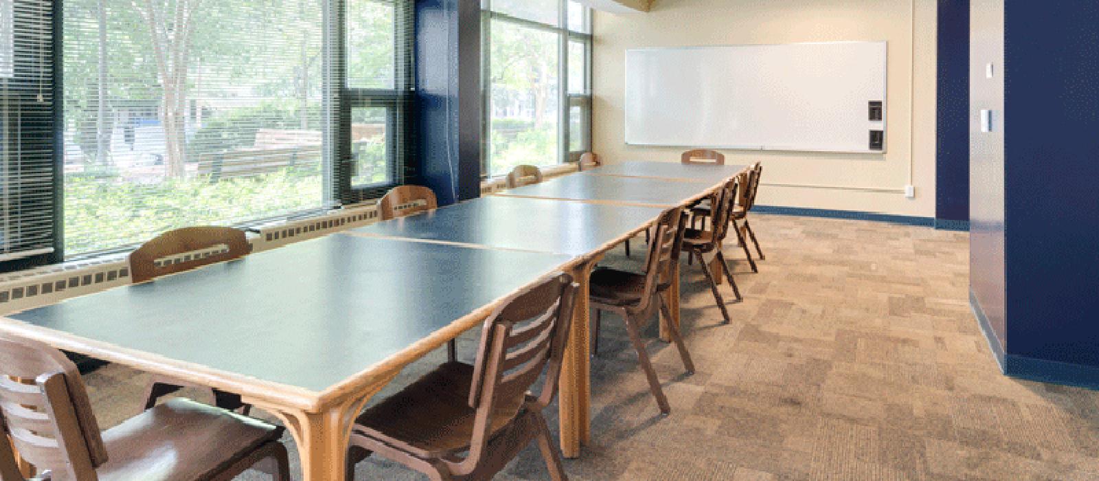 Meeting room with table, chairs, and a white board. One side of the wall is made up of glass pane windows.