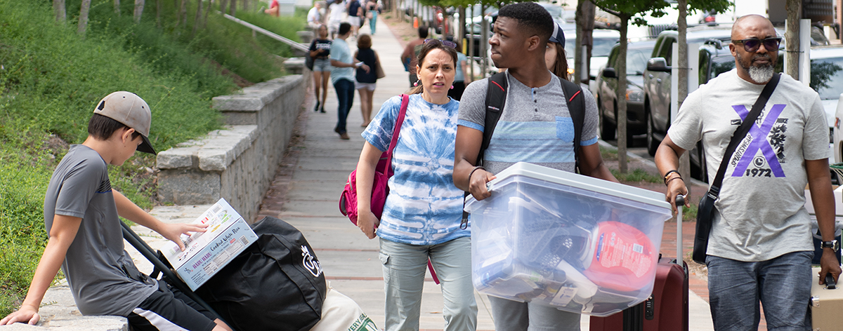 Image of a student carrying a moving bin with his family pulling luggage